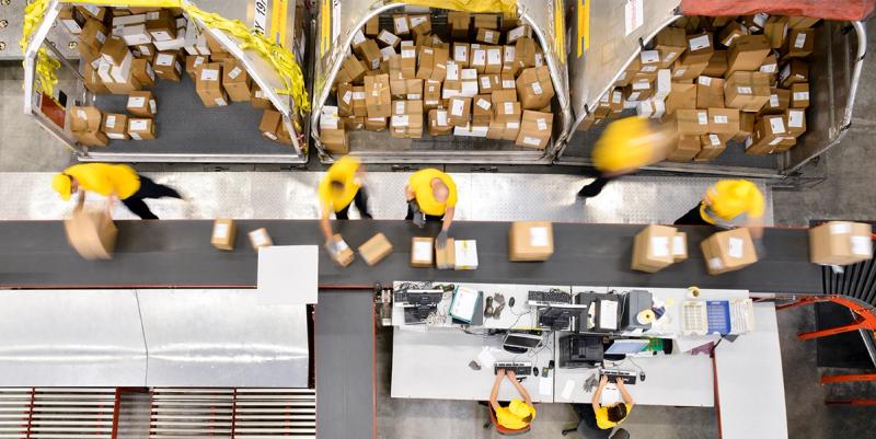 High-angle view of warehouse workers in yellow caps sorting labeled parcels along a conveyor sortation line.