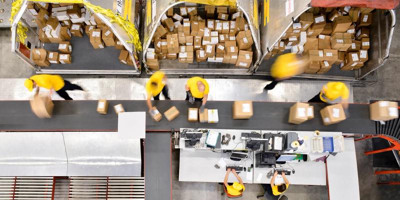 High-angle view of warehouse workers in yellow caps sorting labeled parcels along a conveyor sortation line.