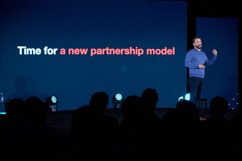 Man on stage before a screen reading 'Time for a new partnership model' with a silhouetted audience
