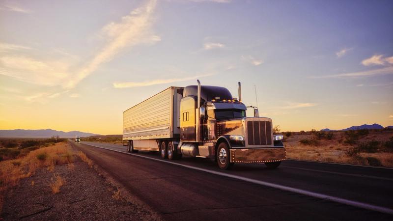 Semi-truck with silver trailer driving down a desert highway at sunset with mountains in the distance