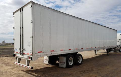 A white enclosed semi-trailer (dry van) with rear swing doors and dual rear axles parked on dirt under a blue sky.