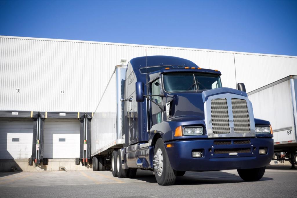 Blue semi truck with trailer backed into a warehouse loading dock under clear blue sky