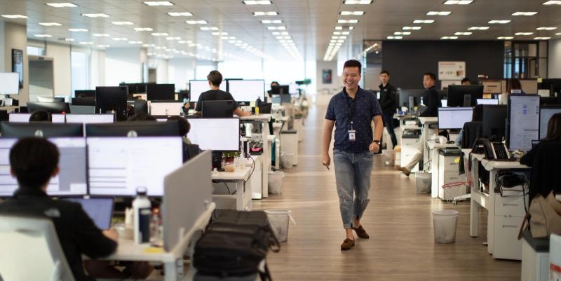 Man in a polo shirt walks smiling down the aisle of a large open-plan office full of working staff.
