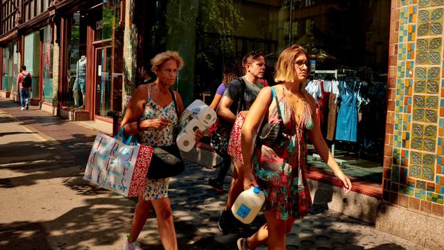 Shoppers walk down a sunny city sidewalk past store windows carrying tote bags, paper towels, and a water jug