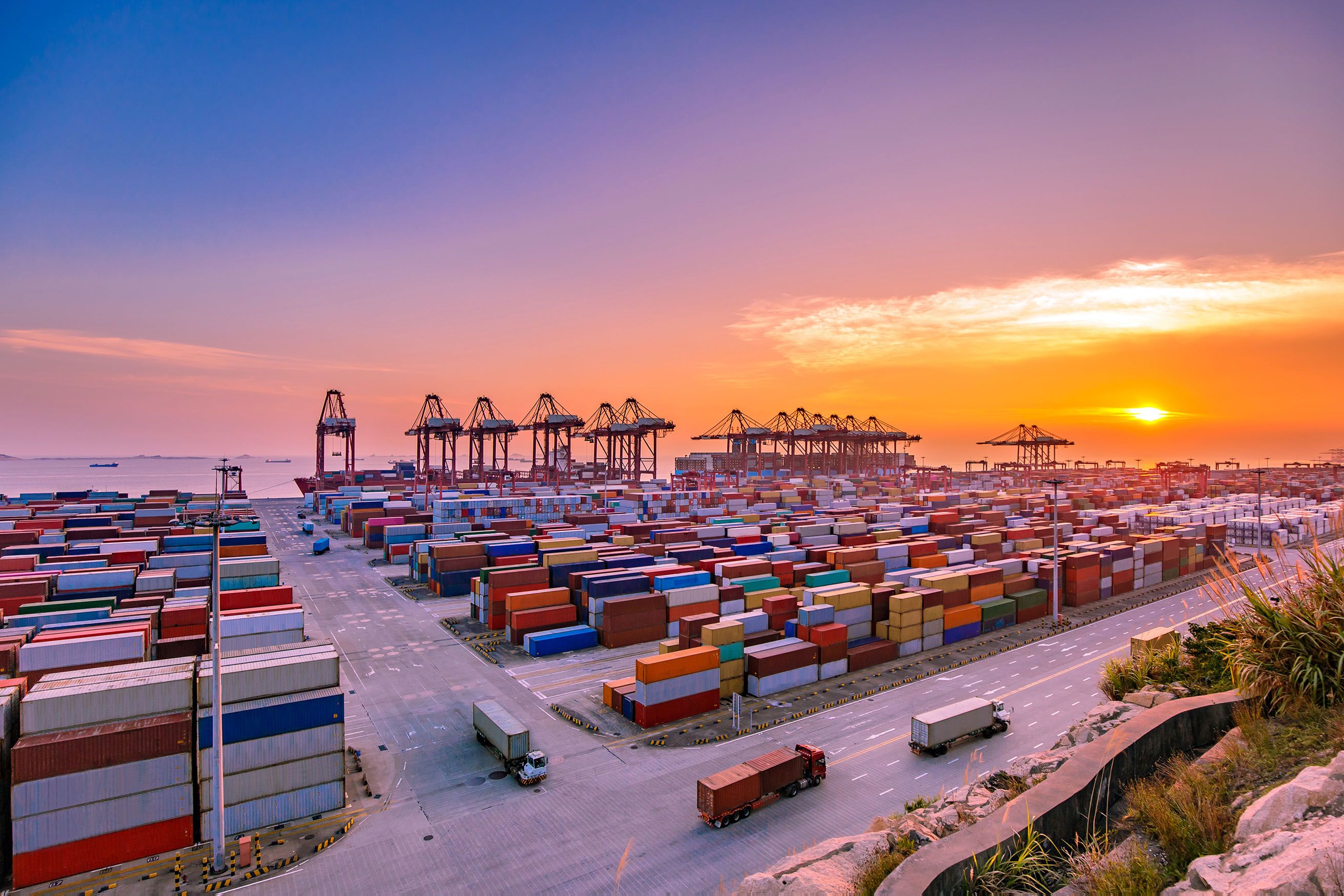 Aerial view of a container port at dusk with stacked shipping containers, gantry cranes, and trucks