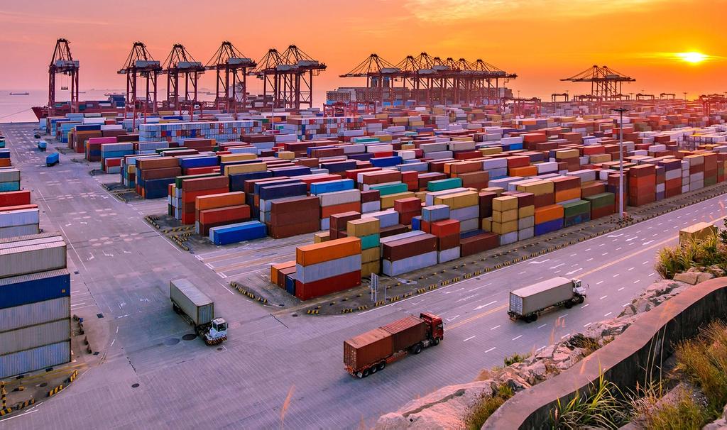 Aerial view of a container port at dusk with stacked shipping containers, gantry cranes, and trucks