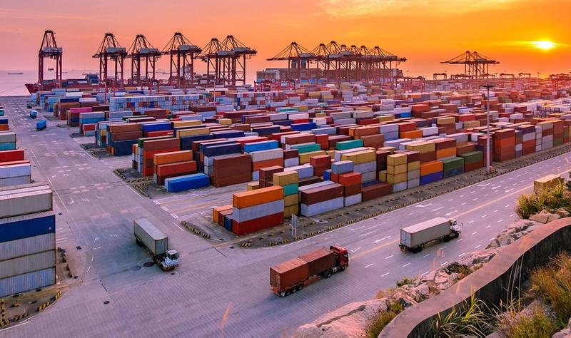 Aerial view of a container port at dusk with stacked shipping containers, gantry cranes, and trucks