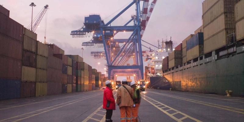 Three workers in hard hats stand at a port terminal at dusk facing gantry cranes and stacked containers