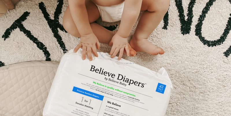 Baby in a diaper crouching over a pack of Believe Diapers on a patterned rug