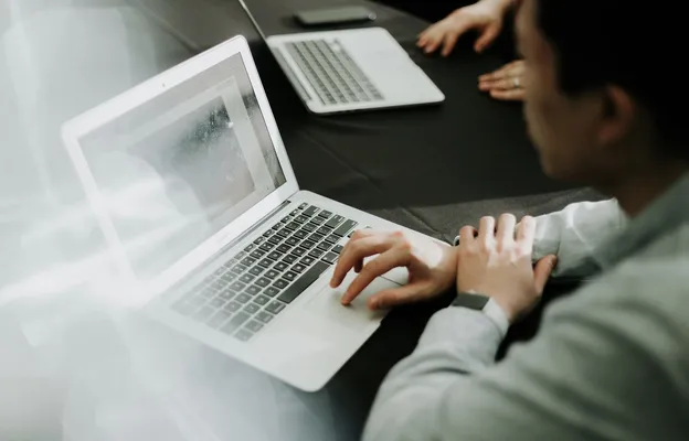 A person in a light-colored shirt is seated at a dark conference table, working on an open laptop with a bright, slightly reflective screen, while another laptop, a smartphone, and colleagues’ hands are visible in the blurred background, suggesting a collaborative meeting or work session.