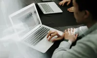 A person in a light-colored shirt is seated at a dark conference table, working on an open laptop with a bright, slightly reflective screen, while another laptop, a smartphone, and colleagues’ hands are visible in the blurred background, suggesting a collaborative meeting or work session.