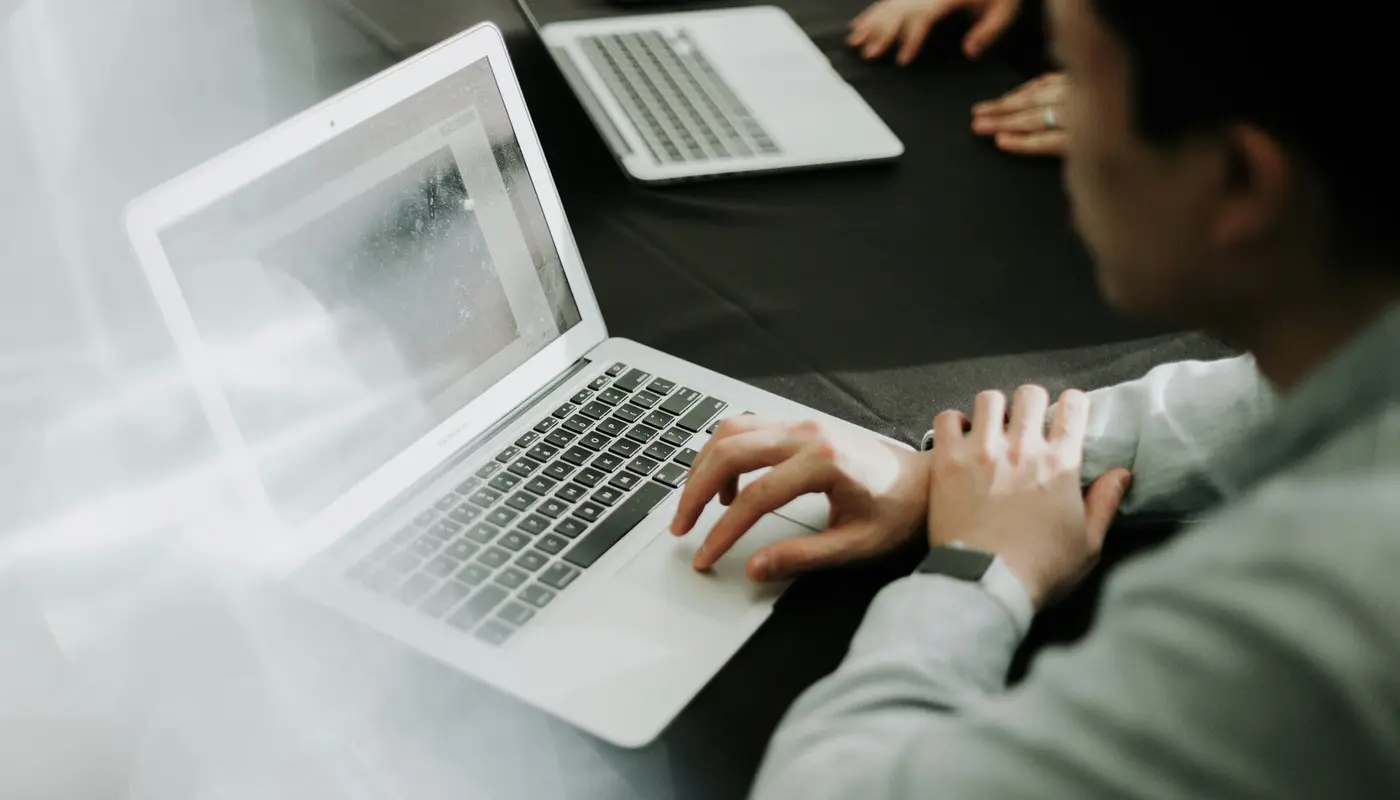 A person in a light-colored shirt is seated at a dark conference table, working on an open laptop with a bright, slightly reflective screen, while another laptop, a smartphone, and colleagues’ hands are visible in the blurred background, suggesting a collaborative meeting or work session.