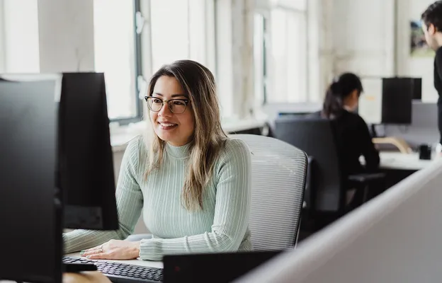 A woman wearing glasses and a light green sweater smiles while working at a desktop computer in a bright office, with another person seated in the background.