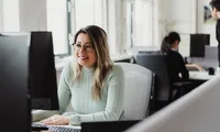 A woman wearing glasses and a light green sweater smiles while working at a desktop computer in a bright office, with another person seated in the background.