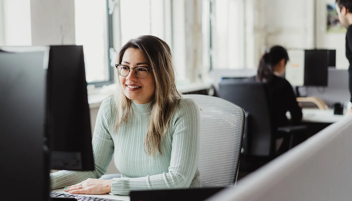 Een vrouw met een bril en een lichtgroene trui glimlacht terwijl ze aan een computer werkt in een lichte kantoorruimte, met een andere persoon op de achtergrond.