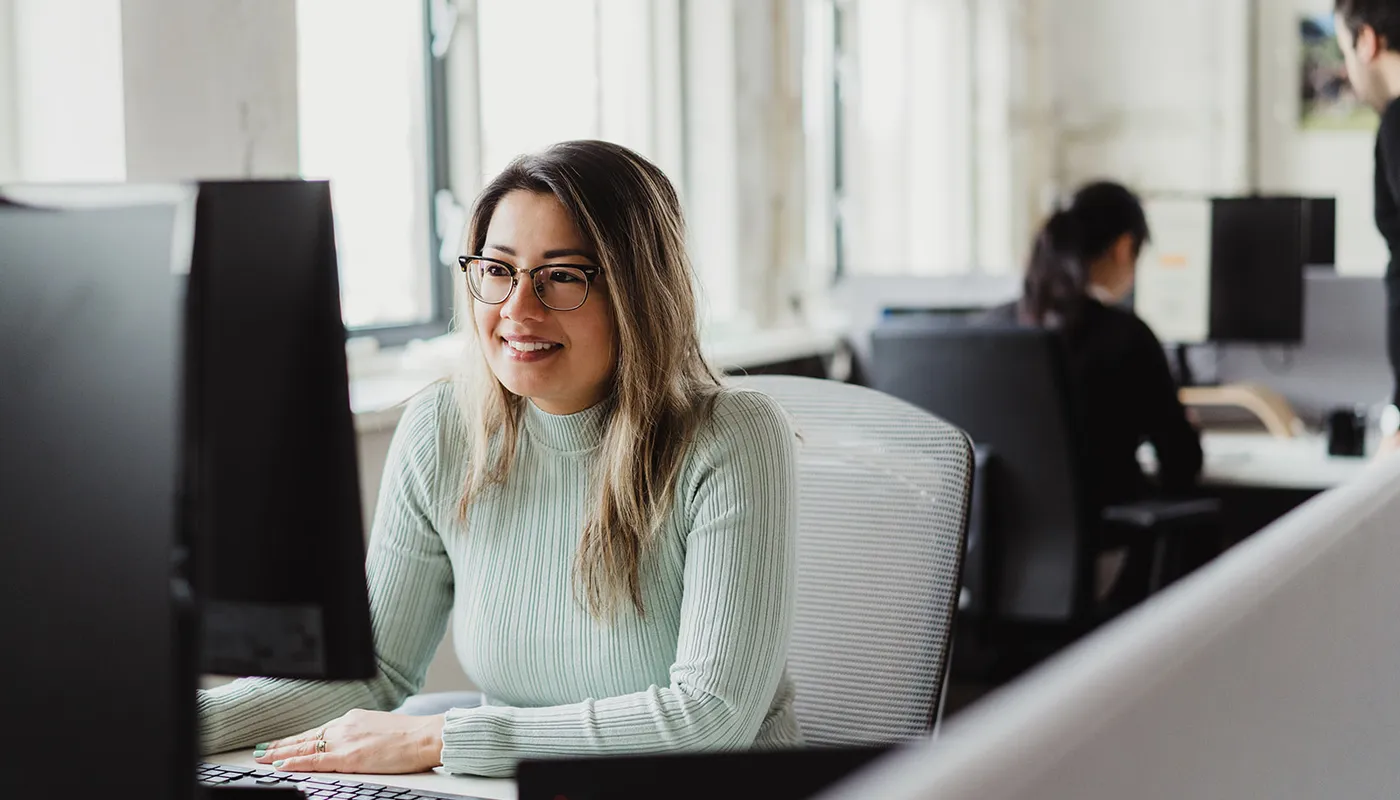 A woman wearing glasses and a light green sweater smiles while working at a desktop computer in a bright office, with another person seated in the background.