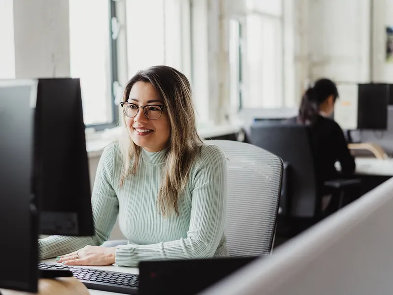 Een vrouw met een bril en een lichtgroene trui glimlacht terwijl ze aan een computer werkt in een lichte kantoorruimte, met een andere persoon op de achtergrond.