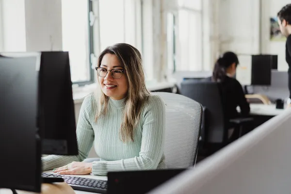 A woman wearing glasses and a light green sweater smiles while working at a desktop computer in a bright office, with another person seated in the background.