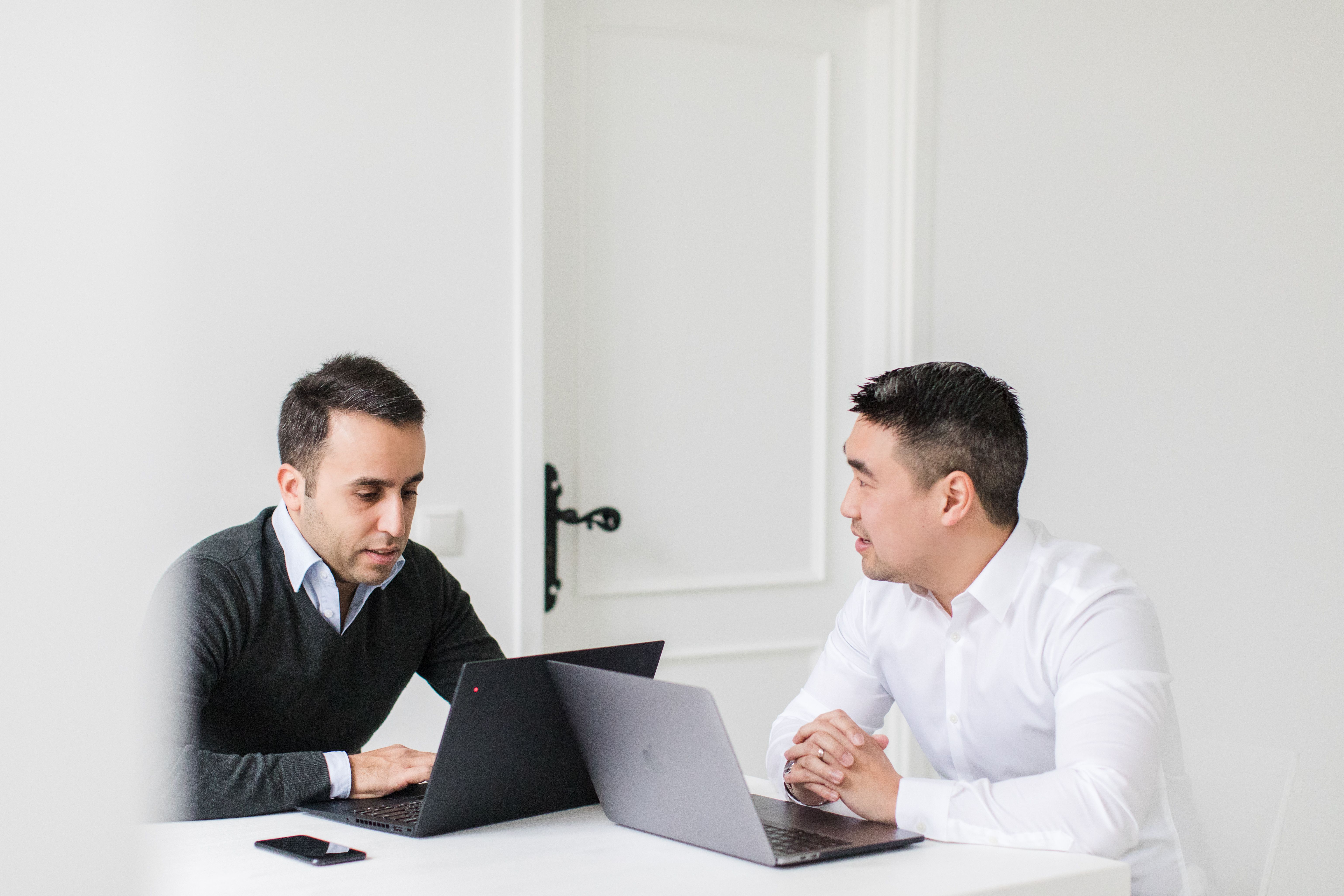 A person in a light-colored shirt is seated at a dark conference table, working on an open laptop with a bright, slightly reflective screen, while another laptop, a smartphone, and colleagues’ hands are visible in the blurred background, suggesting a collaborative meeting or work session.