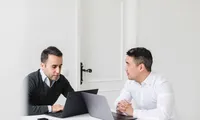 A person in a light-colored shirt is seated at a dark conference table, working on an open laptop with a bright, slightly reflective screen, while another laptop, a smartphone, and colleagues’ hands are visible in the blurred background, suggesting a collaborative meeting or work session.