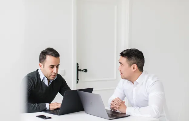 A person in a light-colored shirt is seated at a dark conference table, working on an open laptop with a bright, slightly reflective screen, while another laptop, a smartphone, and colleagues’ hands are visible in the blurred background, suggesting a collaborative meeting or work session.