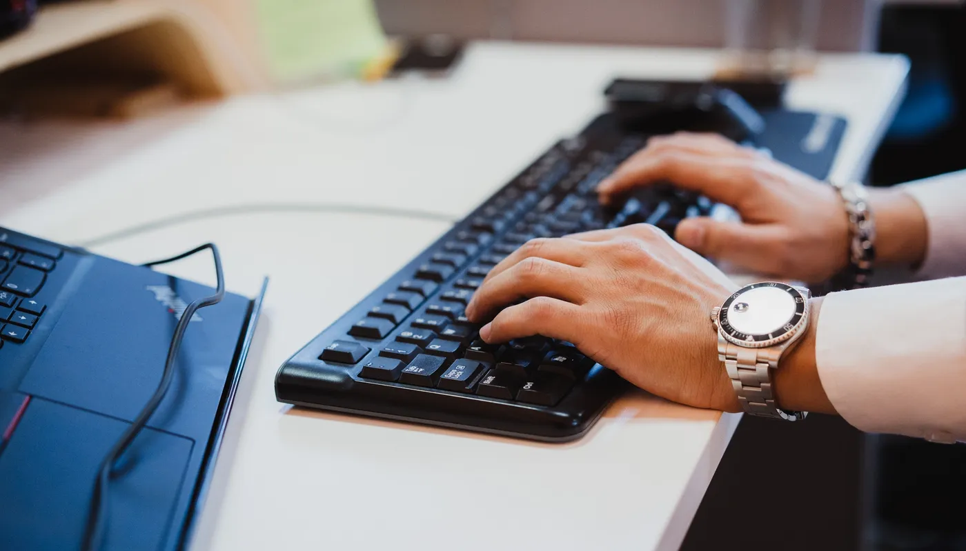 Close-up van een professional die op een toetsenbord typt naast een laptop in een moderne werkruimte, wat productiviteit, technologie en digitale werkomgevingen symboliseert.