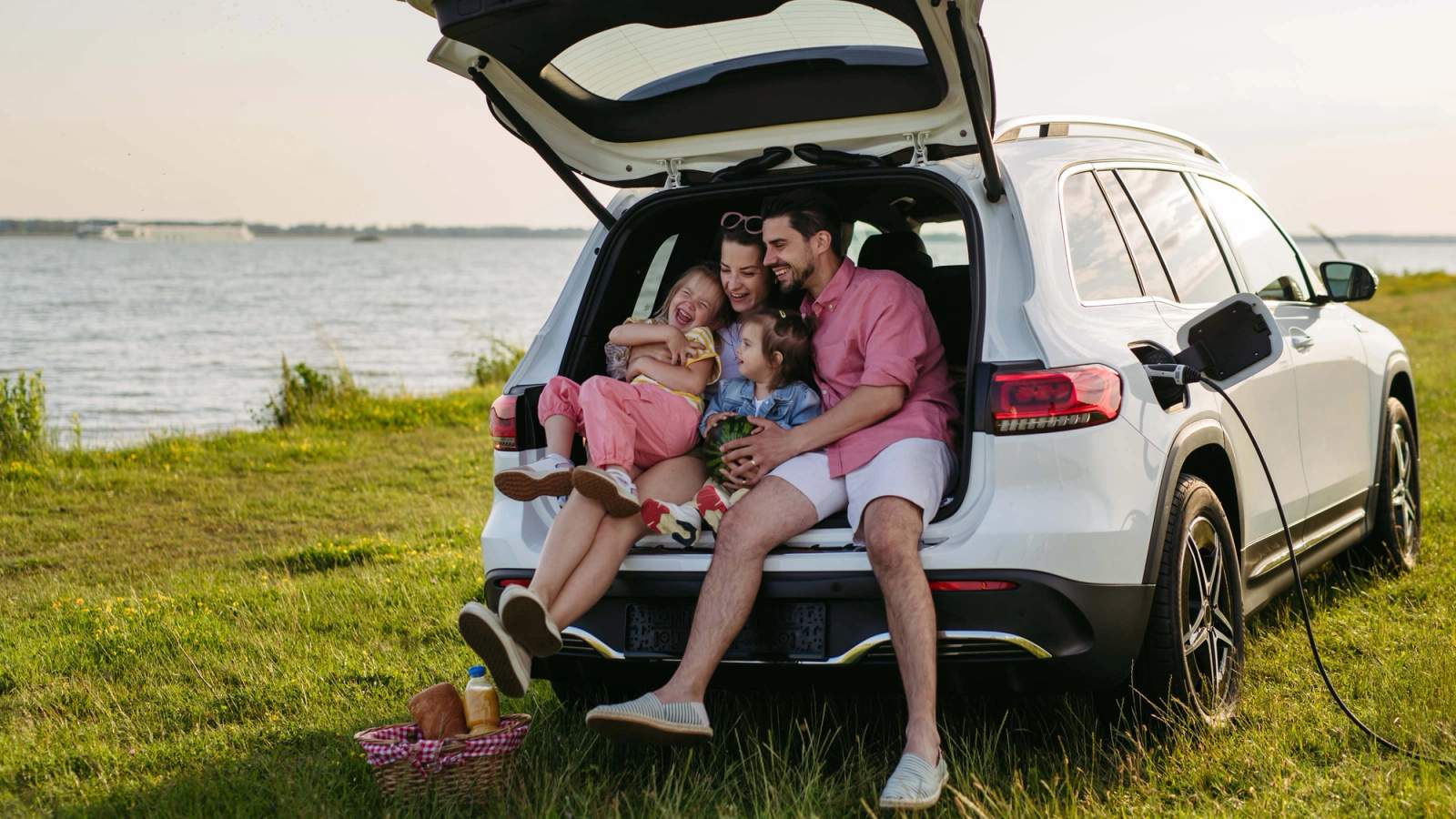 A family sitting in the boot of a Mercedes SUV in summer on grass next to water