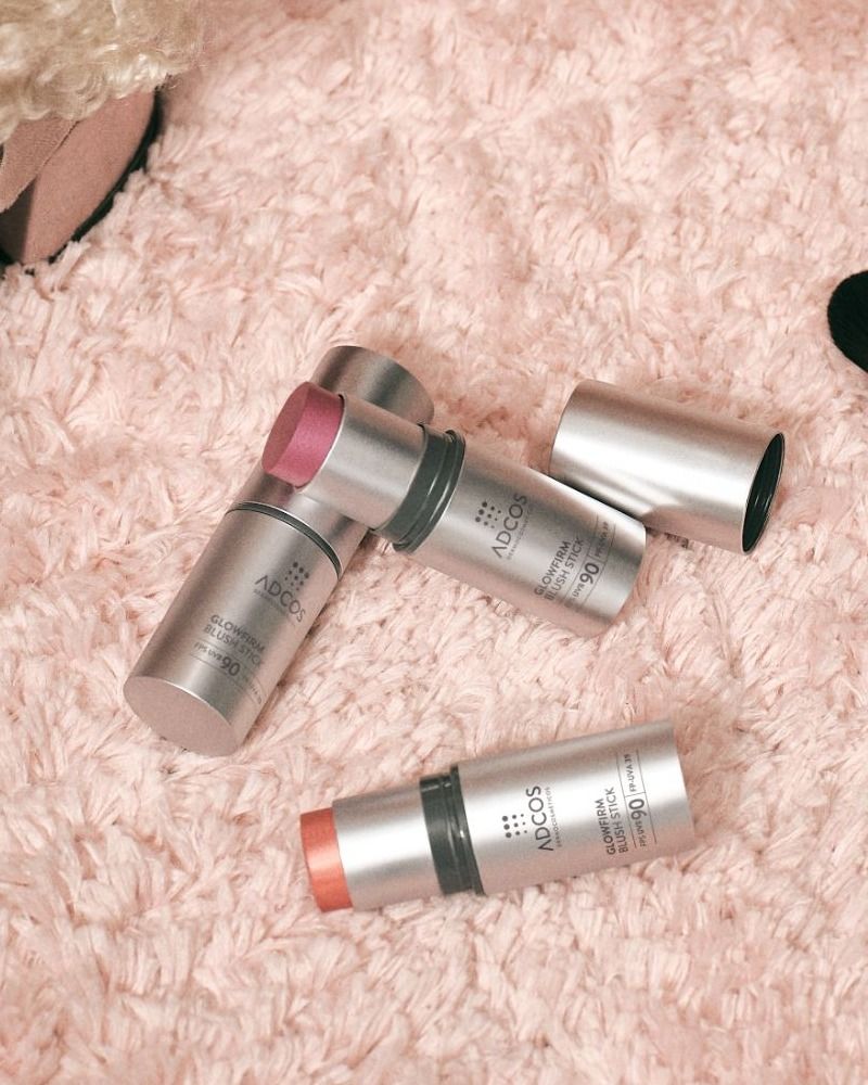 Blush sticks ADCOS Glowfirm sobre tapete rosa, ao lado de sapato e pincéis