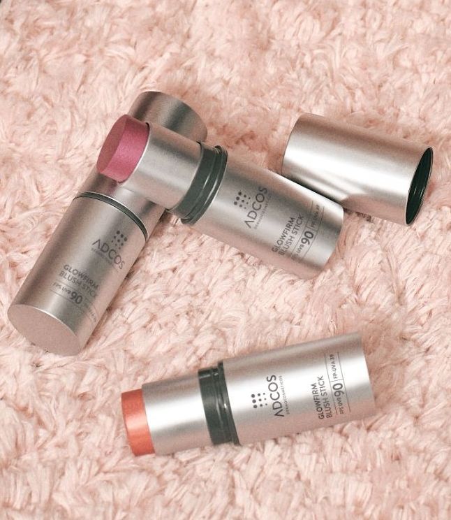 Blush sticks ADCOS Glowfirm sobre tapete rosa, ao lado de sapato e pincéis