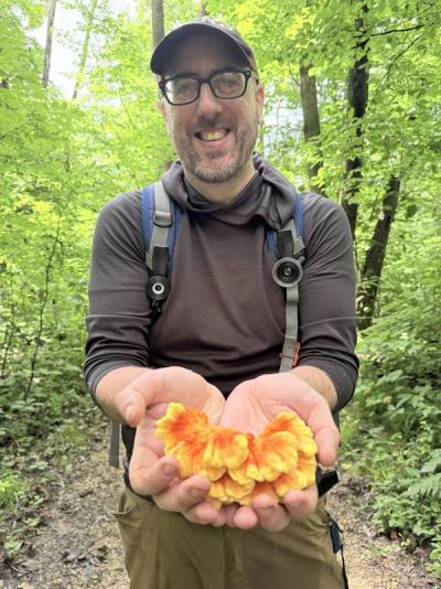 Smiling man with a trimmed beard and glasses holding a small, brightly colored chicken of the woods mushroom up to the camera with both hands.