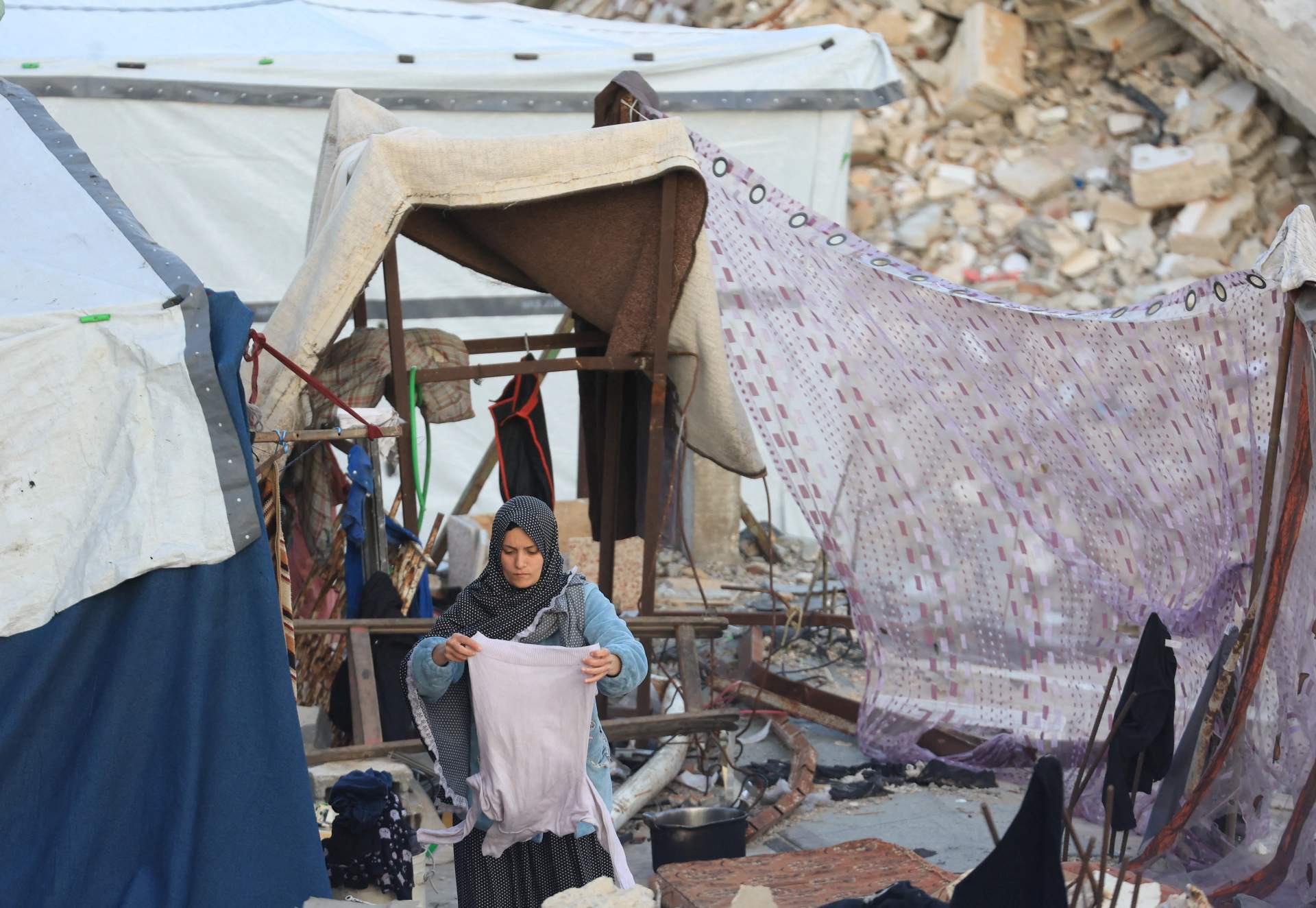 A Palestinian woman in a headscarf holding a shirt while standing outside a makeshift tent amid destroyed buildings in Gaza
