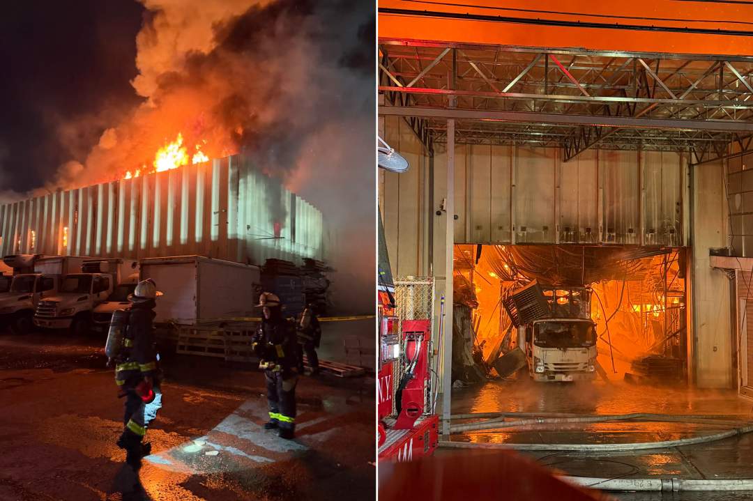 Firefighters in turnout gear and helmets work at night near a large industrial building with emergency lights flashing. Thick smoke is visible in the background as they battle the massive lumberyard fire.