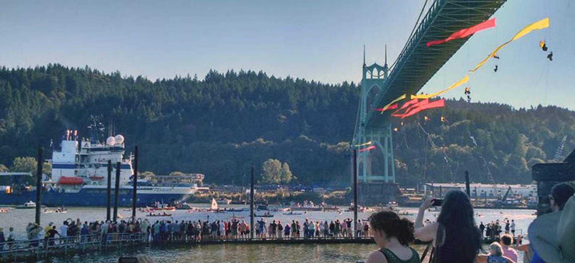 A large crowd lines the waterfront beneath the St. Johns Bridge as the Fennica approaches, with red and yellow banners hanging from the bridge above.