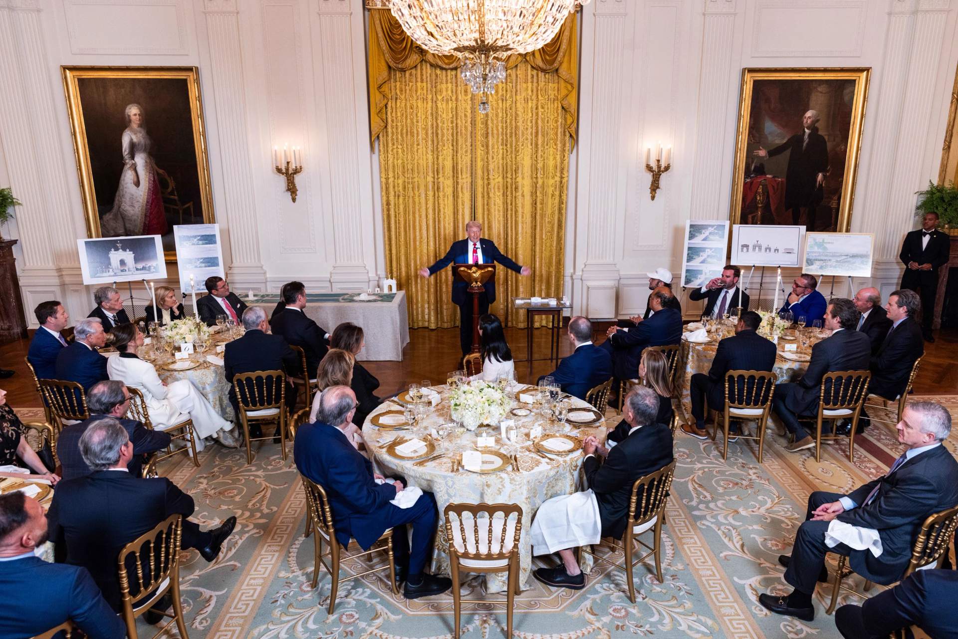 Donald Trump standing at a podium in the decorated East Room of the White House, holding a model of an arch, addressing an audience of corporate donors seated at formal dinner tables