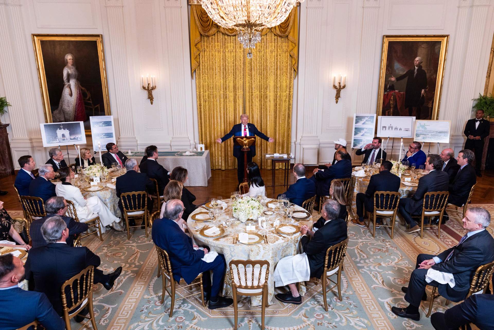 Donald Trump standing at a podium in the decorated East Room of the White House, holding a model of an arch, addressing an audience of corporate donors seated at formal dinner tables