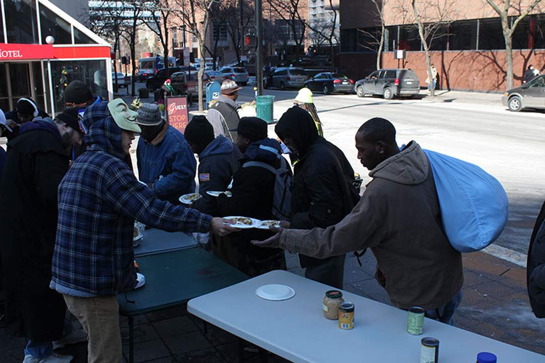 Anonymous activists, one wearing a Guy Fawkes mask, hand out plates of hot food to homeless residents at folding tables set up on a Cincinnati street during an OpSafeWinter event.