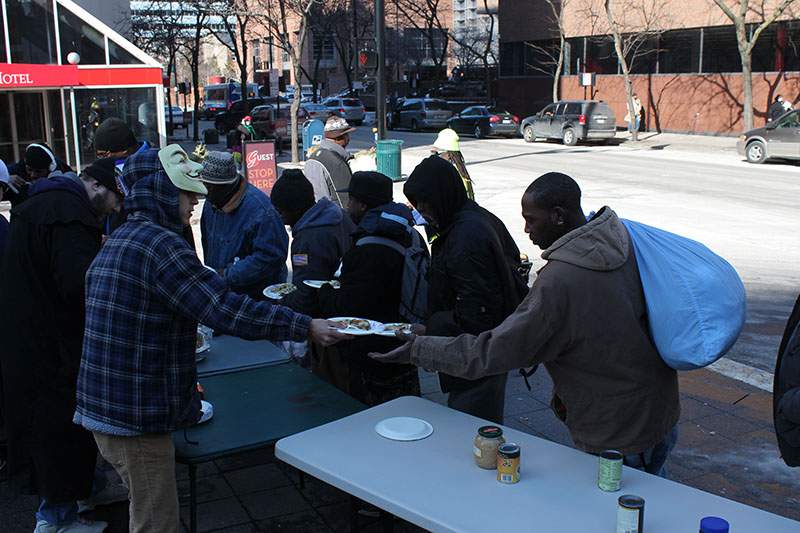 Anonymous activists, one wearing a Guy Fawkes mask, hand out plates of hot food to homeless residents at folding tables set up on a Cincinnati street during an OpSafeWinter event.