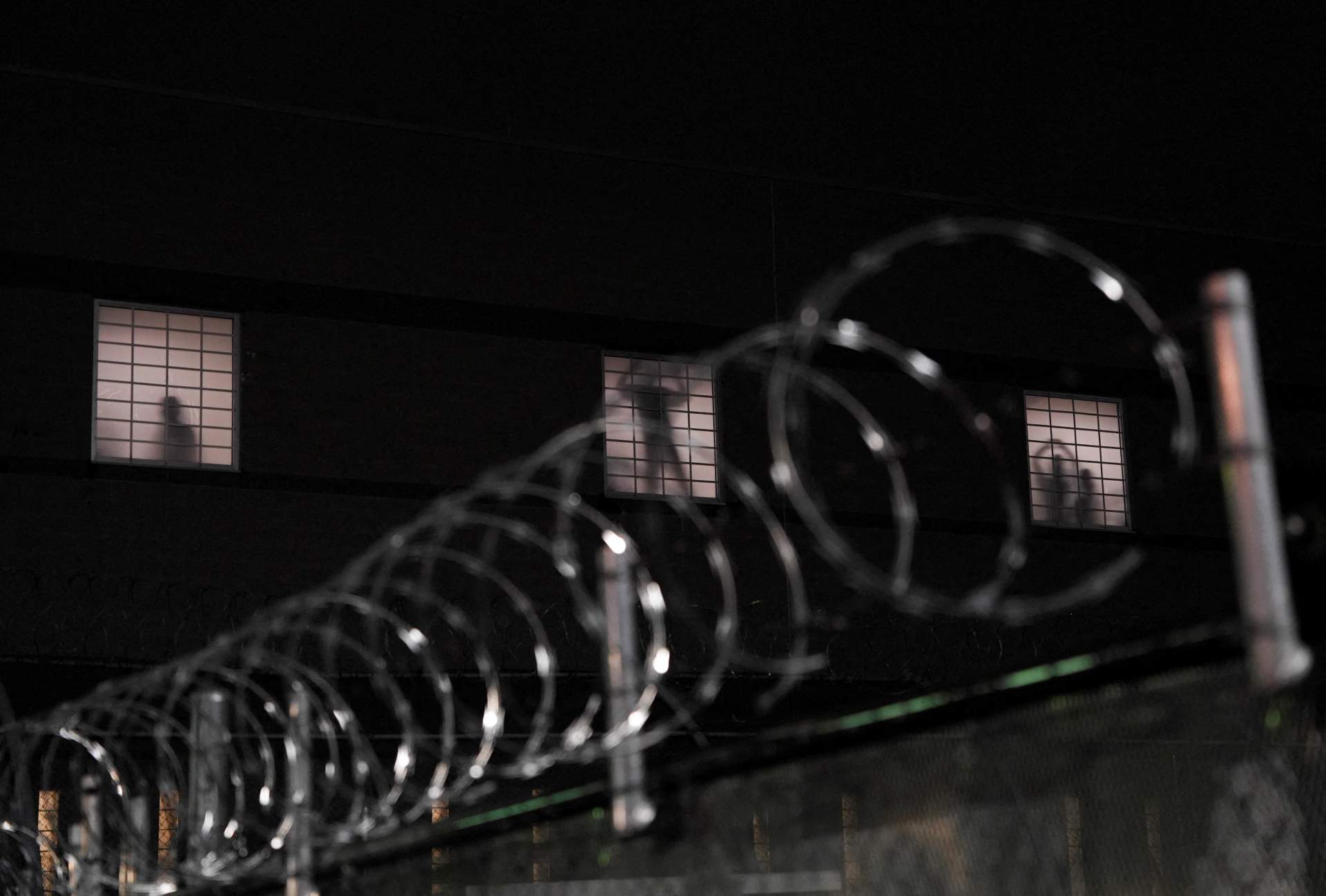 Silhouette of a person visible through a window behind a chain-link fence with razor-wire.