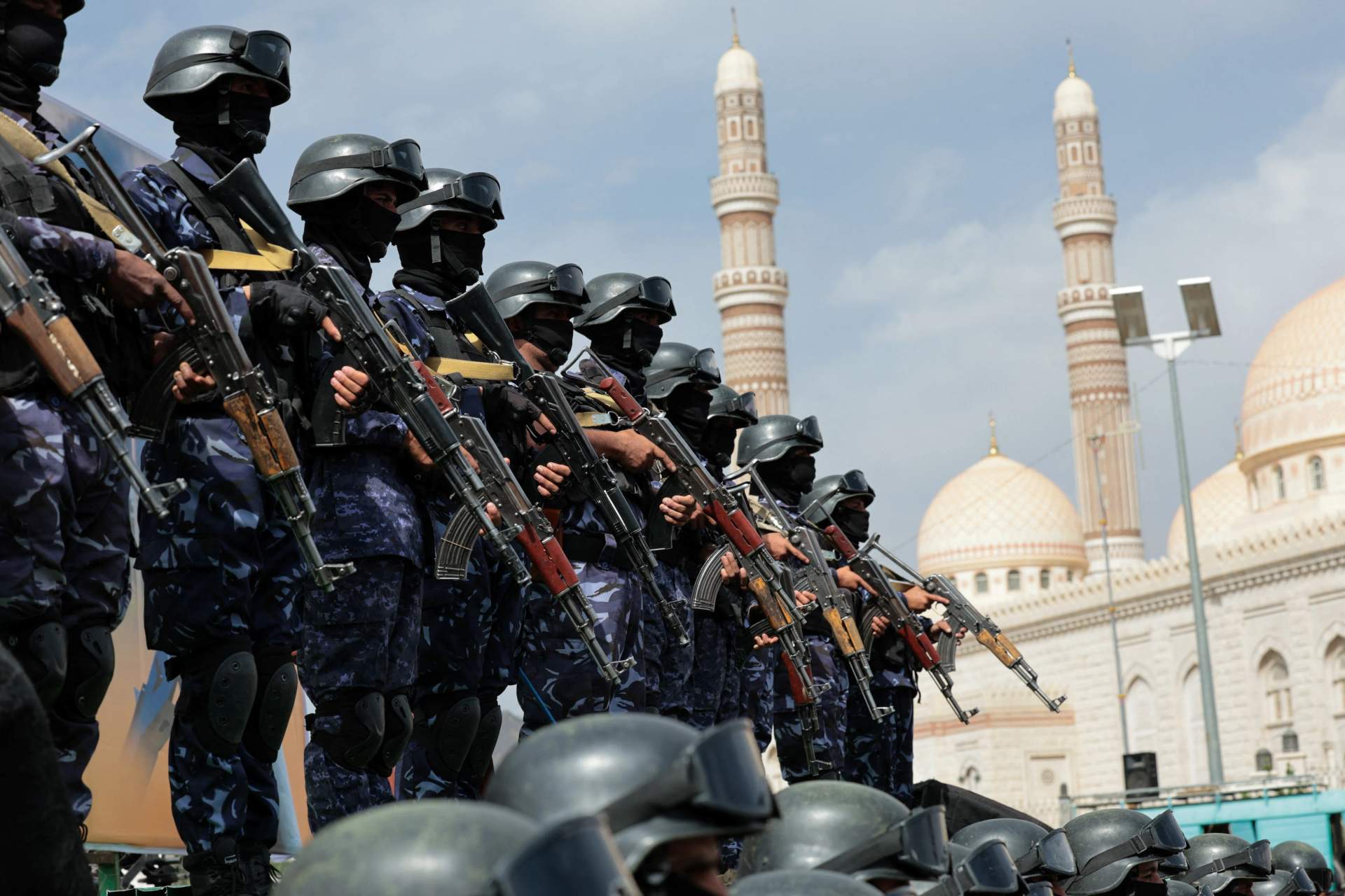 Houthi security personnel in uniform standing in formation at a rally in Sanaa, Yemen, with a crowd visible in the background