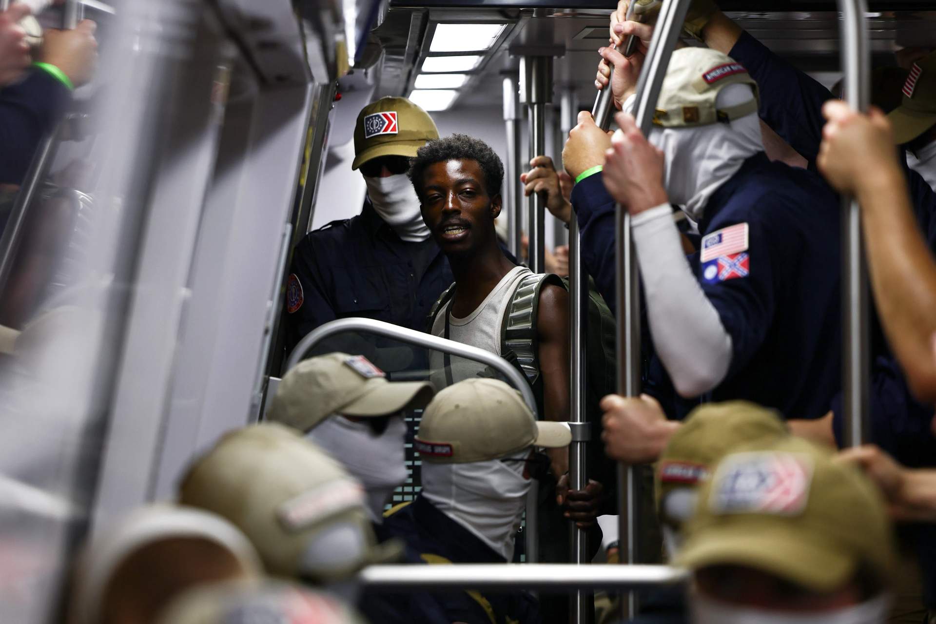 A Black man commuter stands surrounded by masked men in navy blue shirts and khaki pants filling a Metro train car