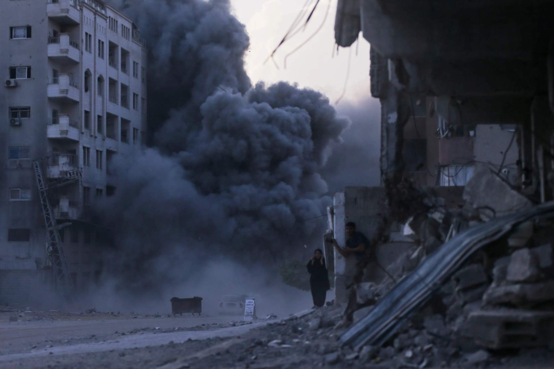 Palestinian civilians running and taking cover on a street in Gaza City during an Israeli airstrike with a large plume of smoke rising from a building