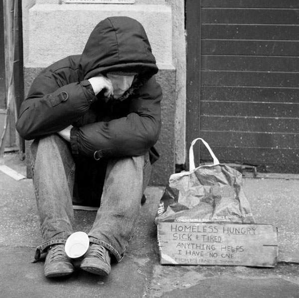 A man sits hunched against a wall next to a cardboard sign reading 'Homeless, hungry, sick and tired. Anything helps. I have no one.'