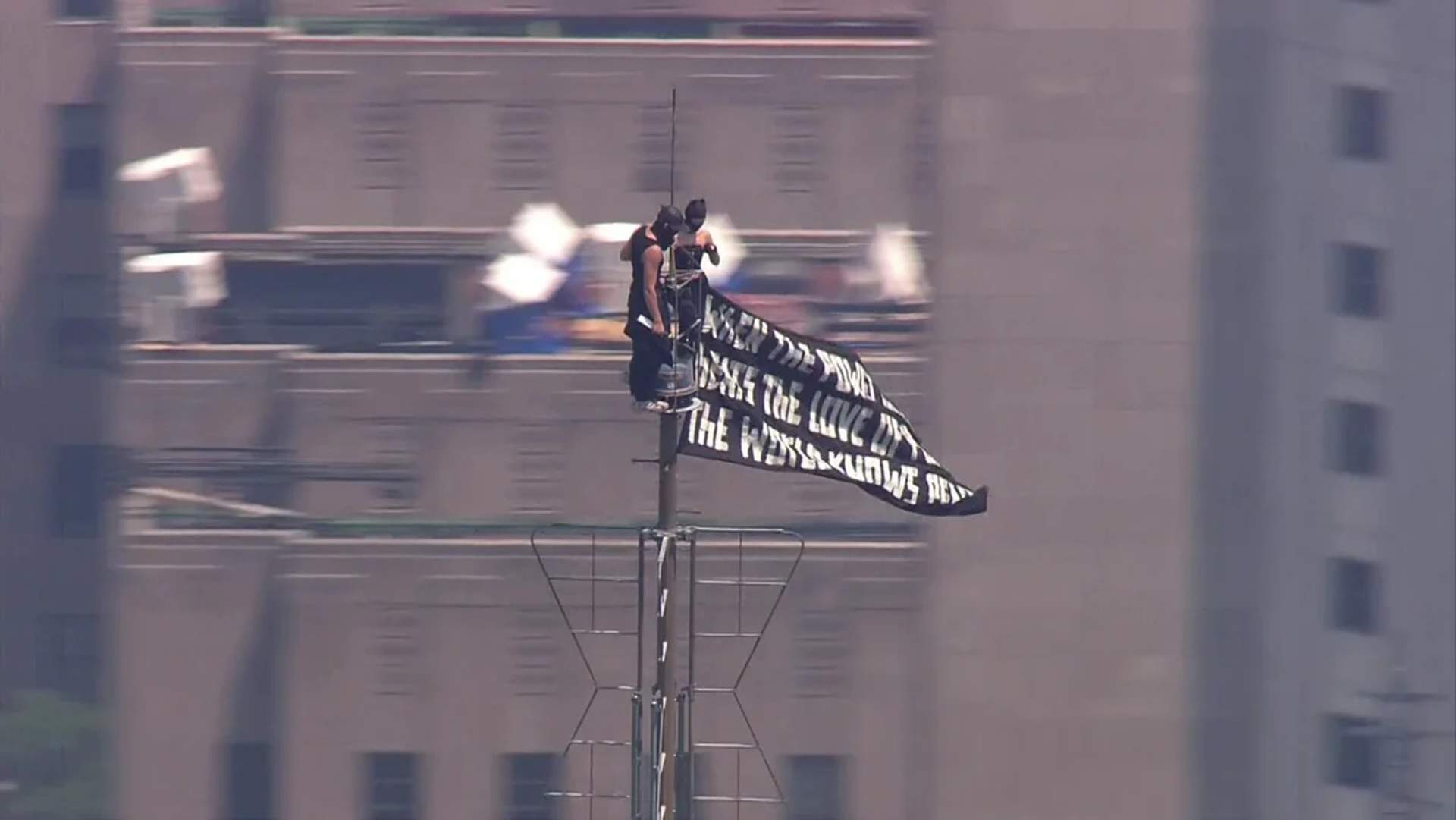 Two masked climbers perch on the Empire State Building antenna spire, holding a black-and-white peace banner, 1,454 feet above Midtown Manhattan on July 1, 2026.