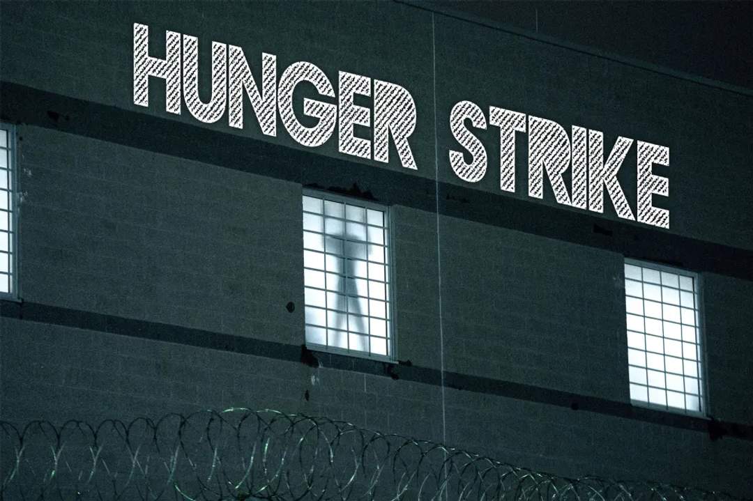 Silhouette of a person visible through a window behind a razor-wire fence, with HUNGER STRIKE projected above him.