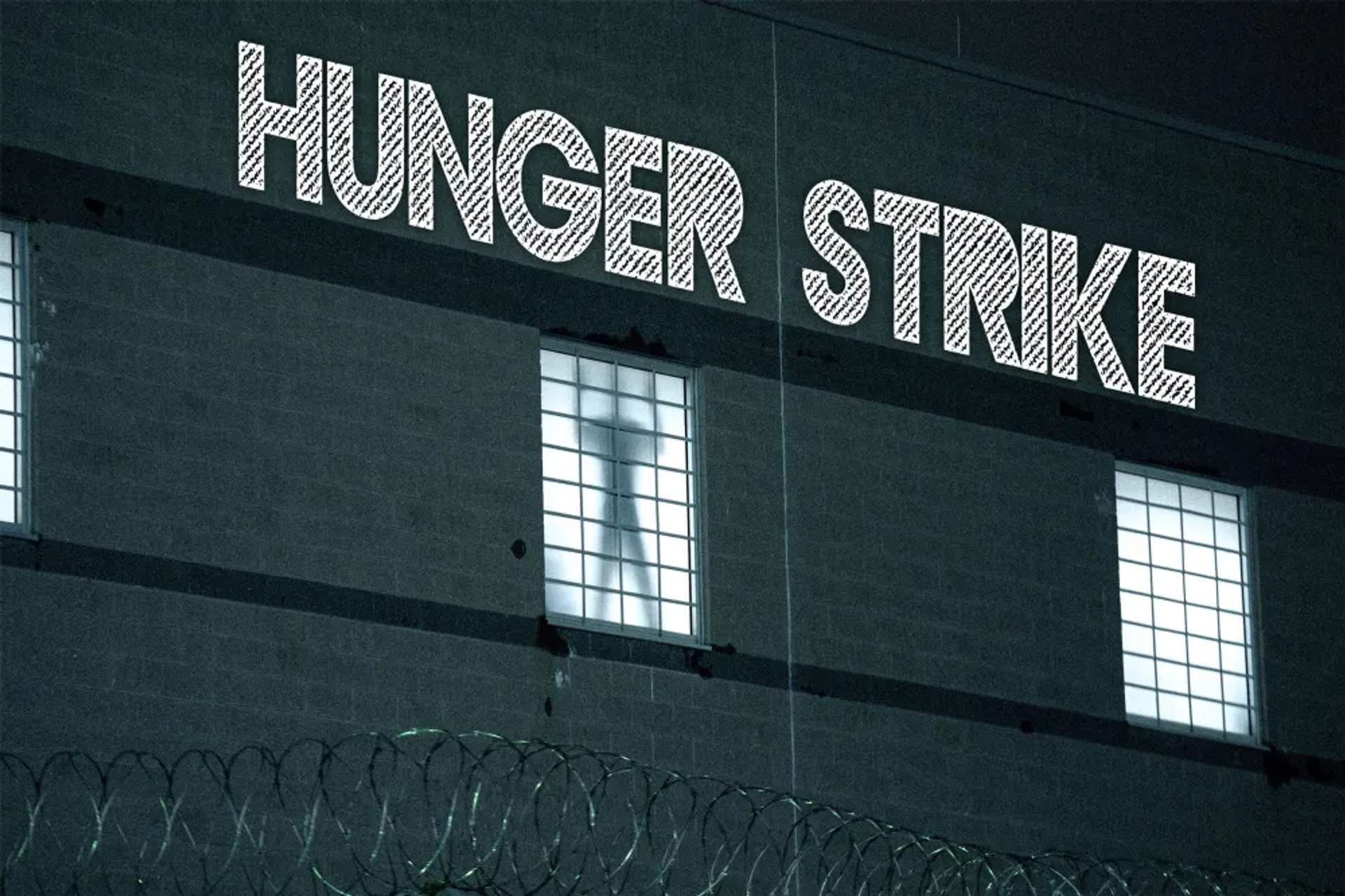 Silhouette of a person visible through a window behind a razor-wire fence, with HUNGER STRIKE projected above him.