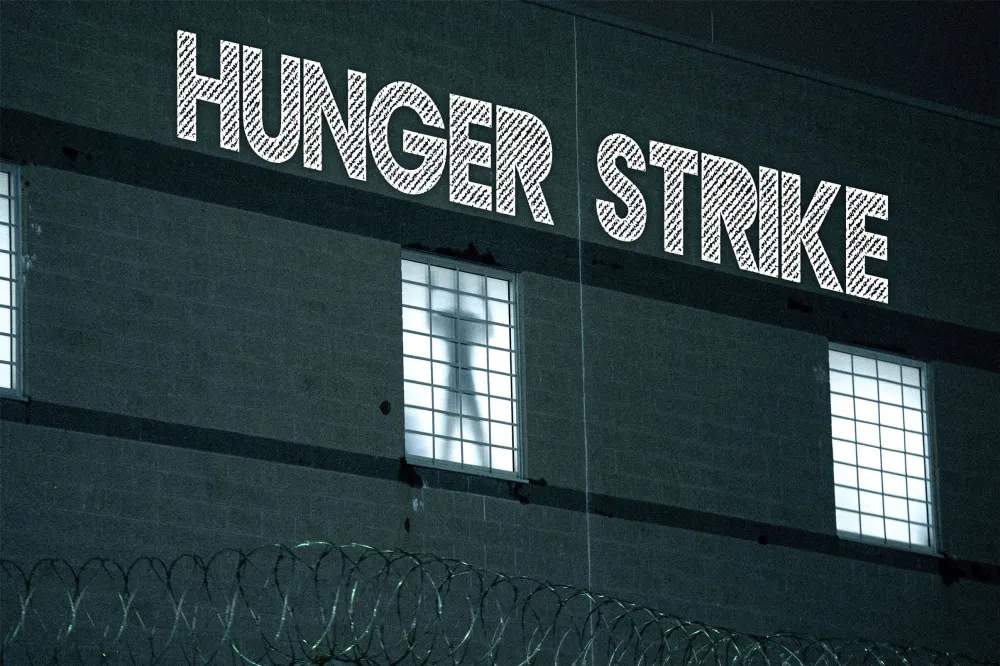 Silhouette of a person visible through a window behind a razor-wire fence, with HUNGER STRIKE projected above him.