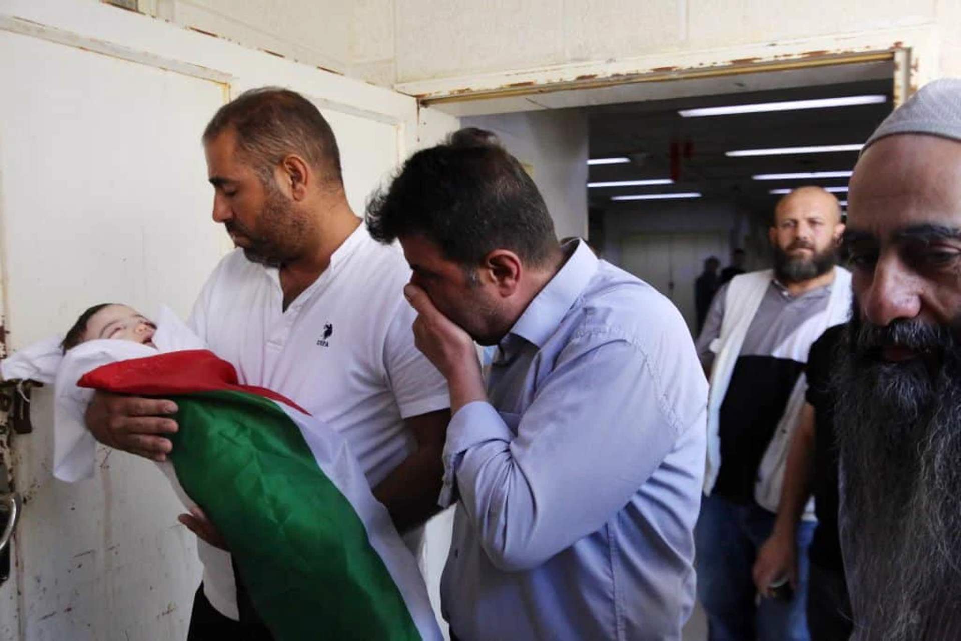 Pallbearers carrying a small wrapped body through narrow streets surrounded by Palestinian mourners