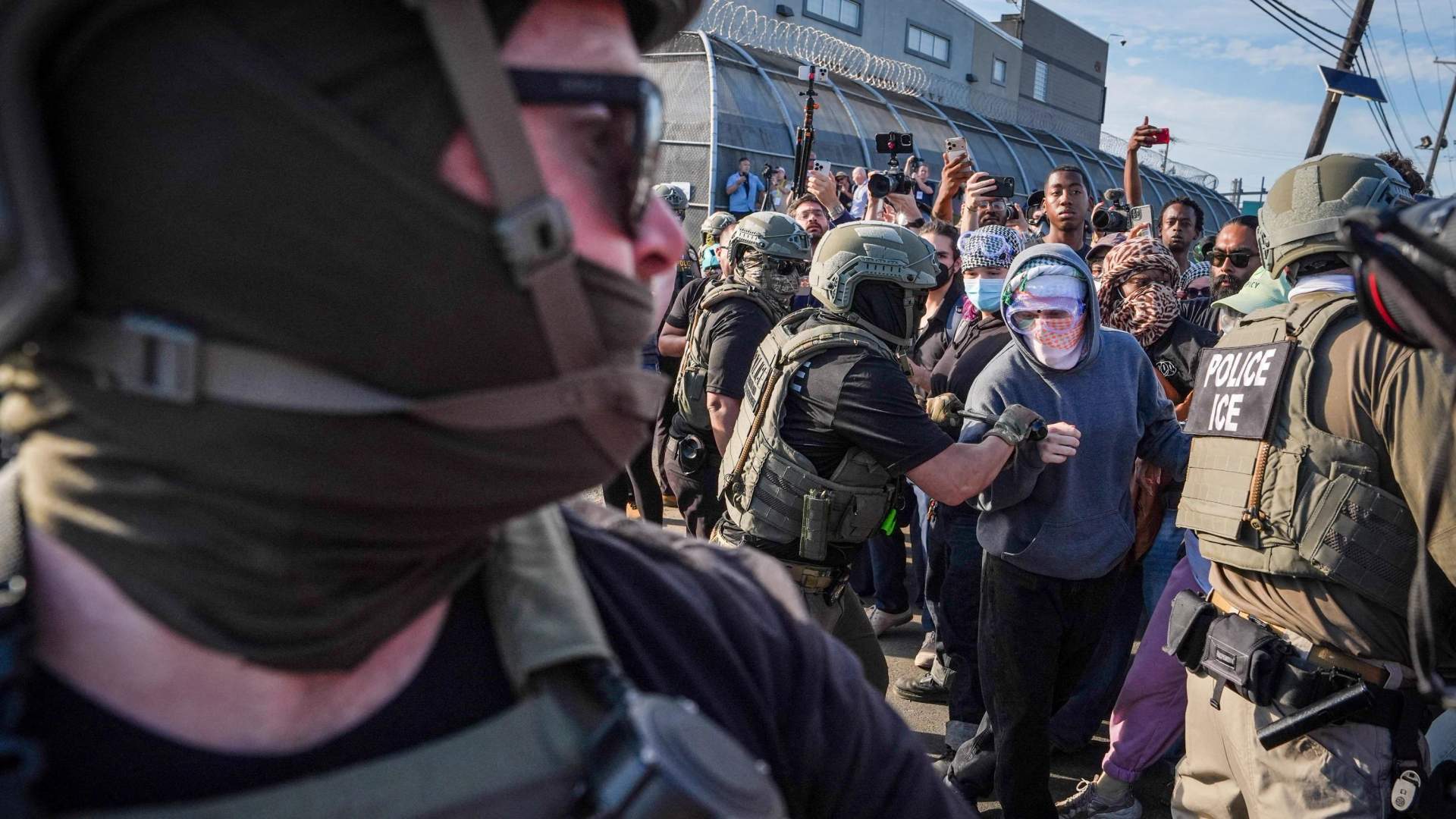Protesters with signs and flags facing ICE agents in tactical gear outside a detention facility