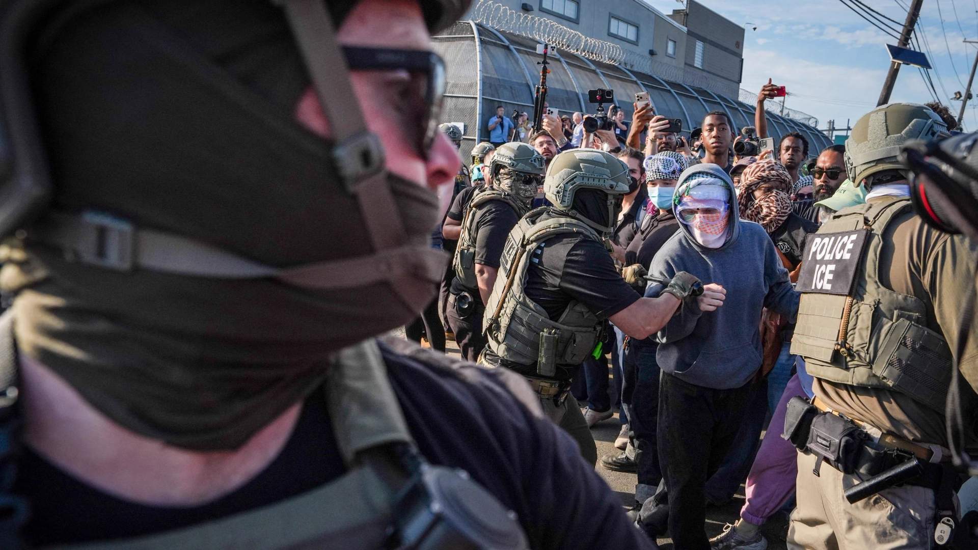 Protesters with signs and flags facing ICE agents in tactical gear outside a detention facility