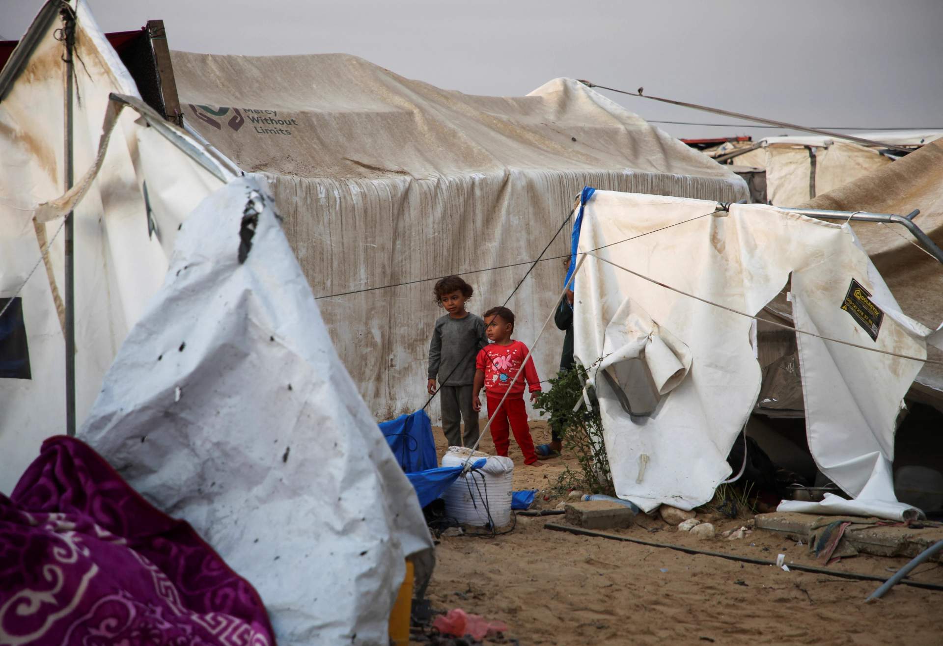 Destroyed tent camp with mangled canvas shelters, debris, and personal belongings scattered on sandy ground after an Israeli airstrike on displaced Palestinians in Gaza